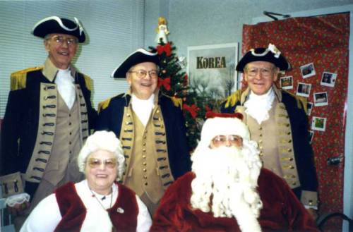 Shawnee Mission Sertoma Club members at a Veterans Administration hospital. The three members stand in a room behind people dressed as Santa Claus and Mrs. Santa Claus, who are seated. The members wear Revolutionary War uniforms with tri corner hats. Mrs. Claus wears a red vest, white blouse, and white wig. Santa wears a plush red velvet jacket, long white beard, and red and white hat. A Christmas tree is behind the group. A door at the right is covered with Christmas gift wrapping. A poster near it has the title: ""KOREA"". A window covered with a venetian blind is at the left. 